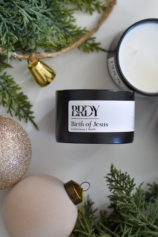 Black candle jar labeled 'Birth of Jesus' with Christmas decorations on a light background