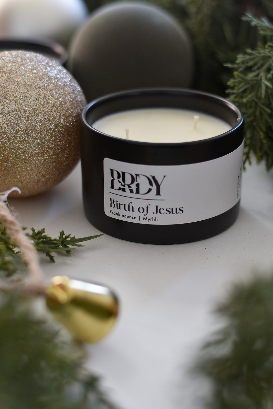 Candle in a black container with 'BDRY' label on a white surface with Christmas decorations.
