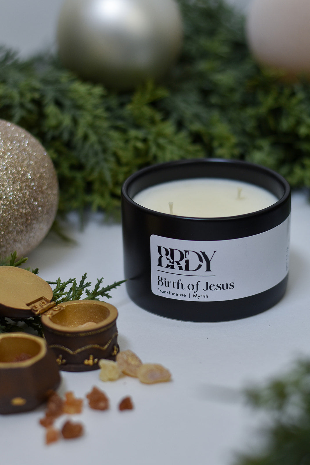 Black candle container that says "BIrth of Jesus" christmas candle with branding on a white surface with greenery and gold ornaments.
